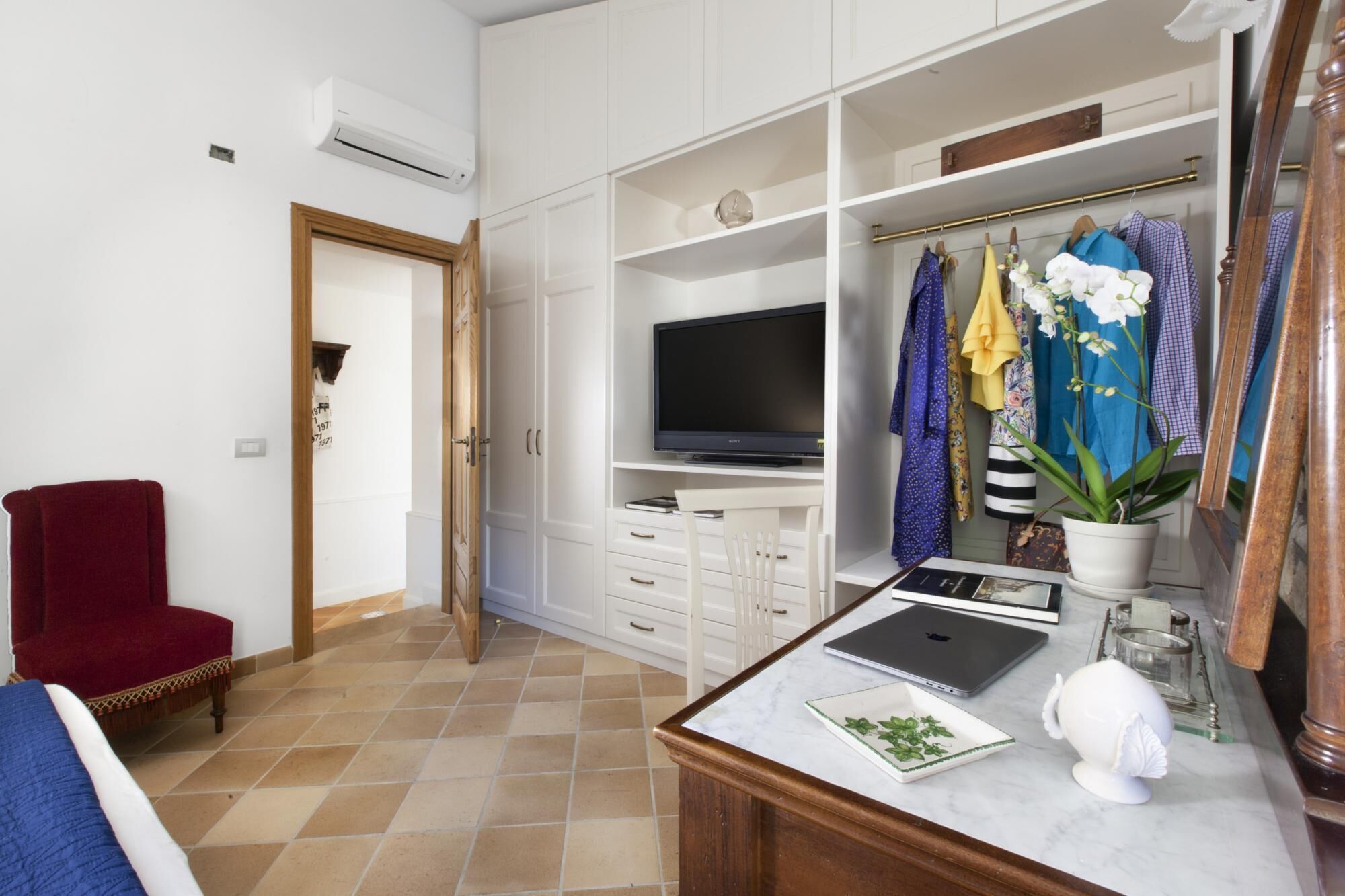 A well-organized room with a television, a desk with a laptop, a chair, and a wardrobe with hanging clothes.