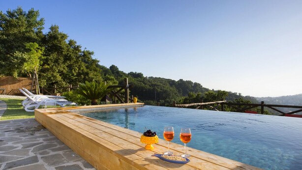 An infinity pool with a wooden deck, two glasses of red wine, and a small yellow fruit bowl.