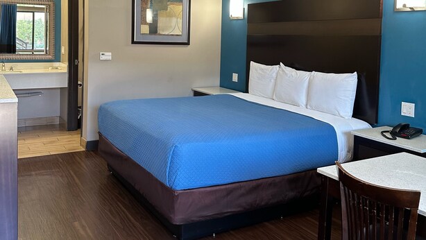 A hotel room with a large bed, a desk, a chair, a framed picture on the wall, and a window with curtains. - Crystal Palace Inn (Maywood)