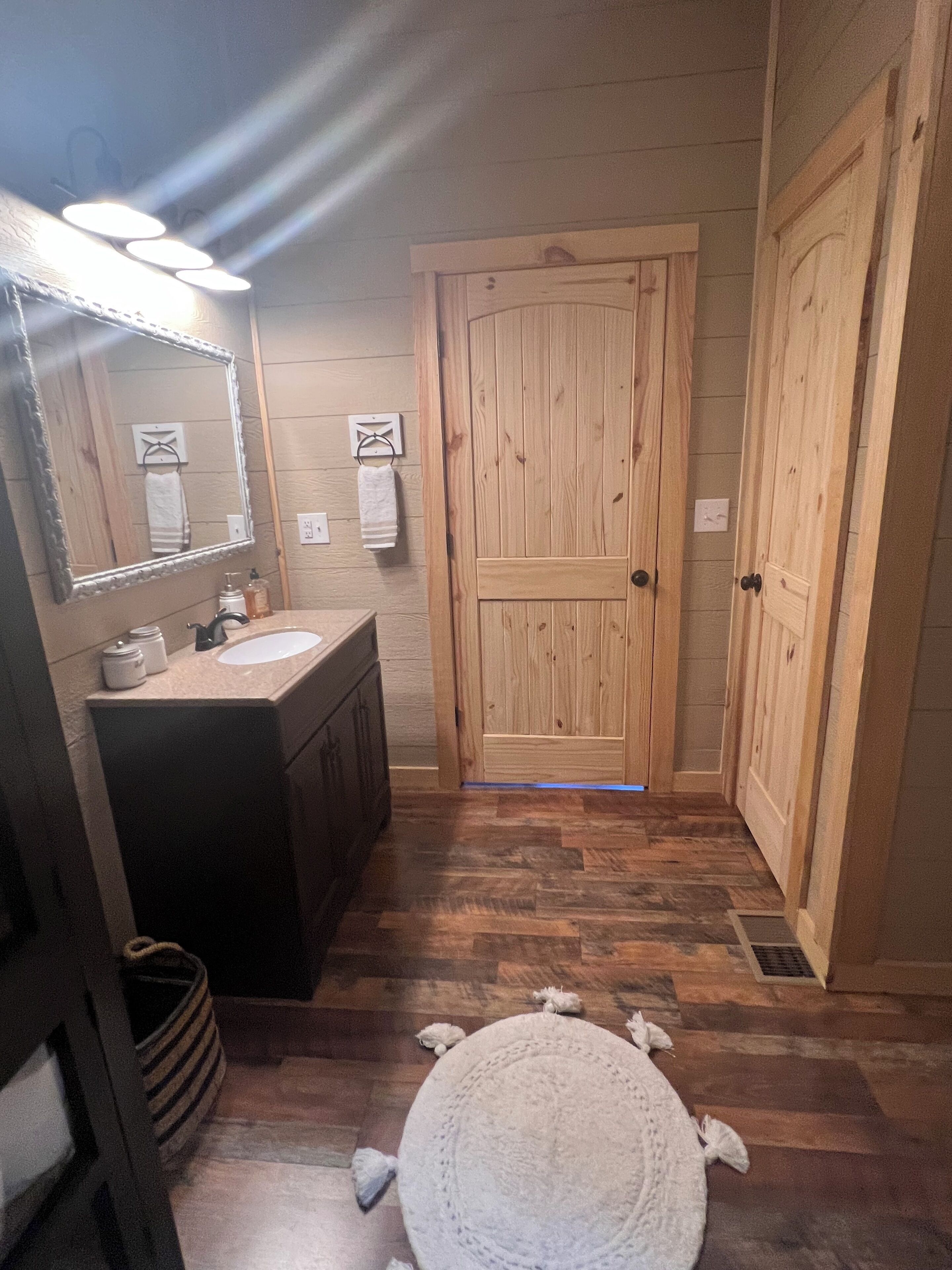 A bathroom with a wooden door, a sink with a mirror, and a round rug on the floor.