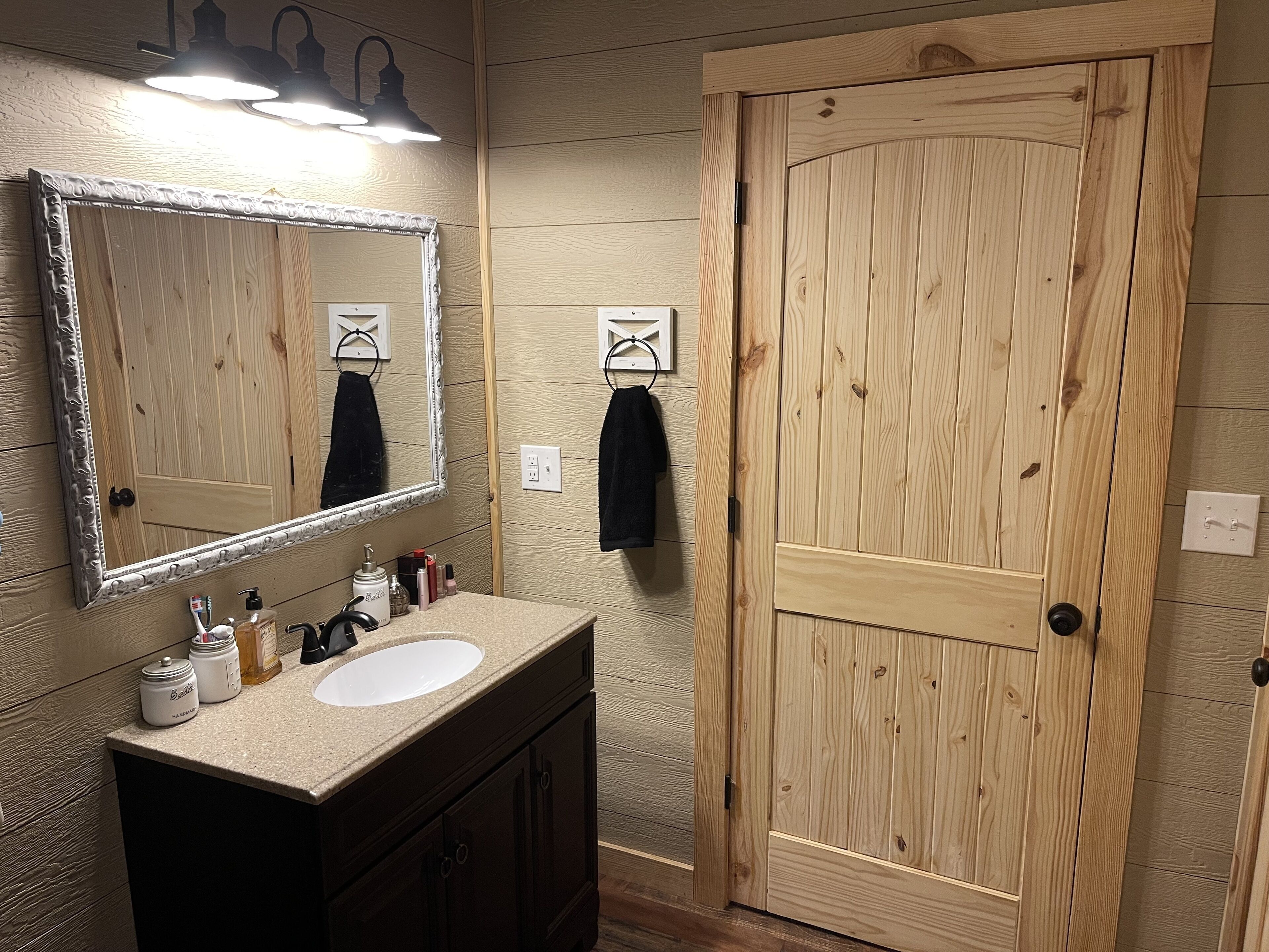 A bathroom with a wooden door, a large mirror, and a vanity with toiletries.
