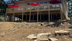 Una terraza de madera de dos pisos con sombrillas rojas, rodeada de árboles y hierba seca.