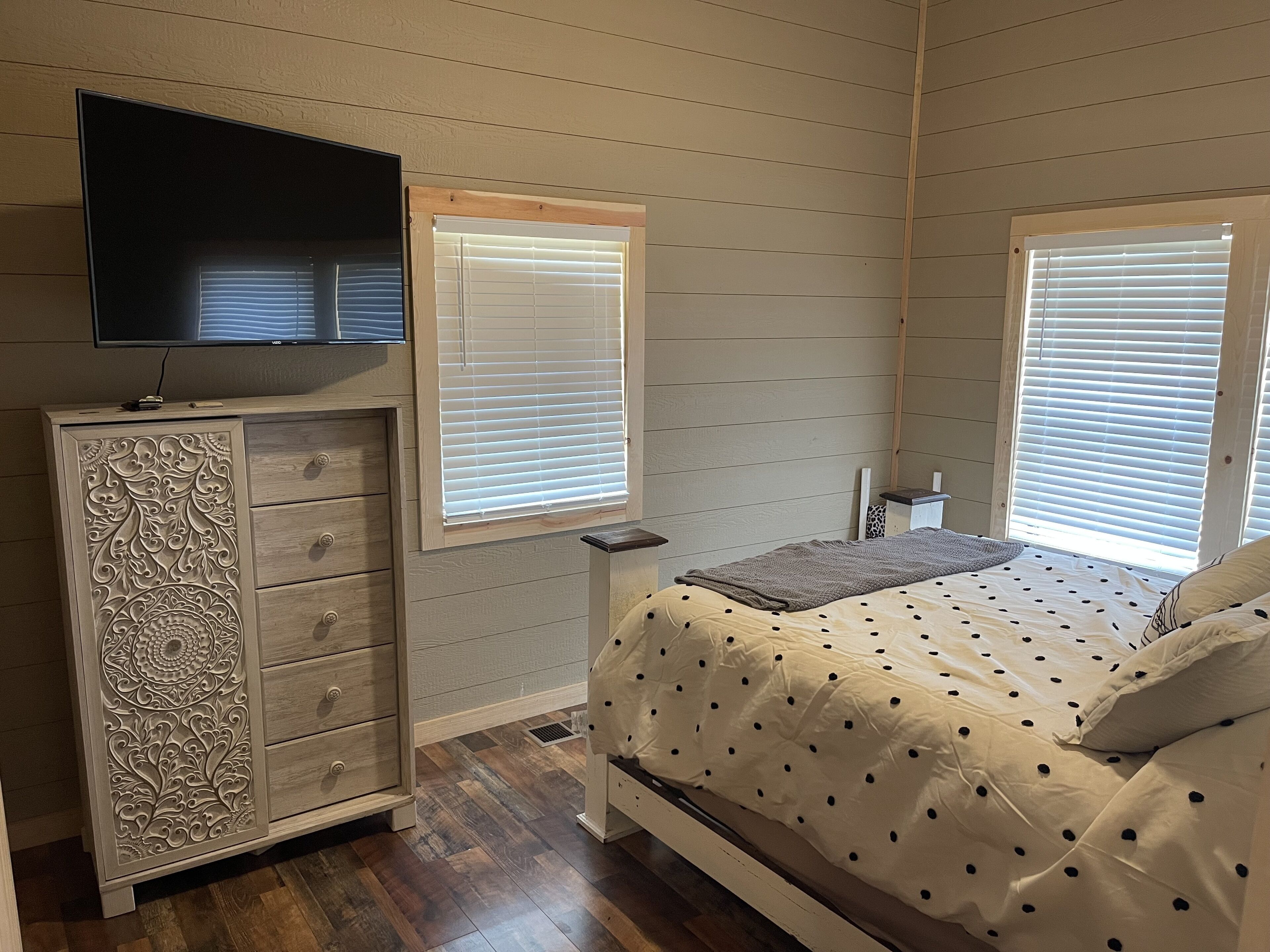 A bedroom with a bed, dresser, TV, and two windows with blinds.