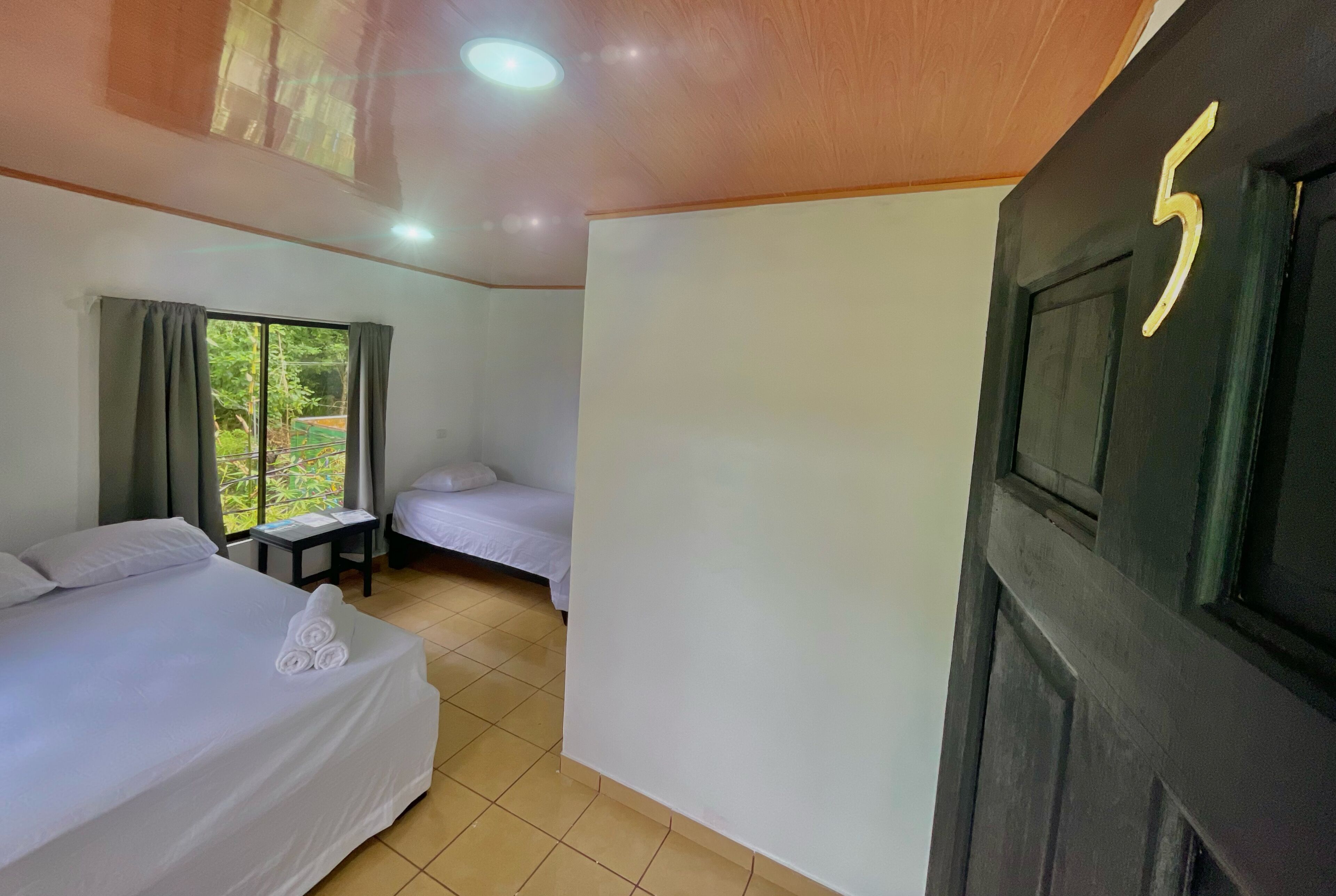 Habitación de hotel con dos camas, una mesita, un ventanal con cortinas y una puerta con el número 5.