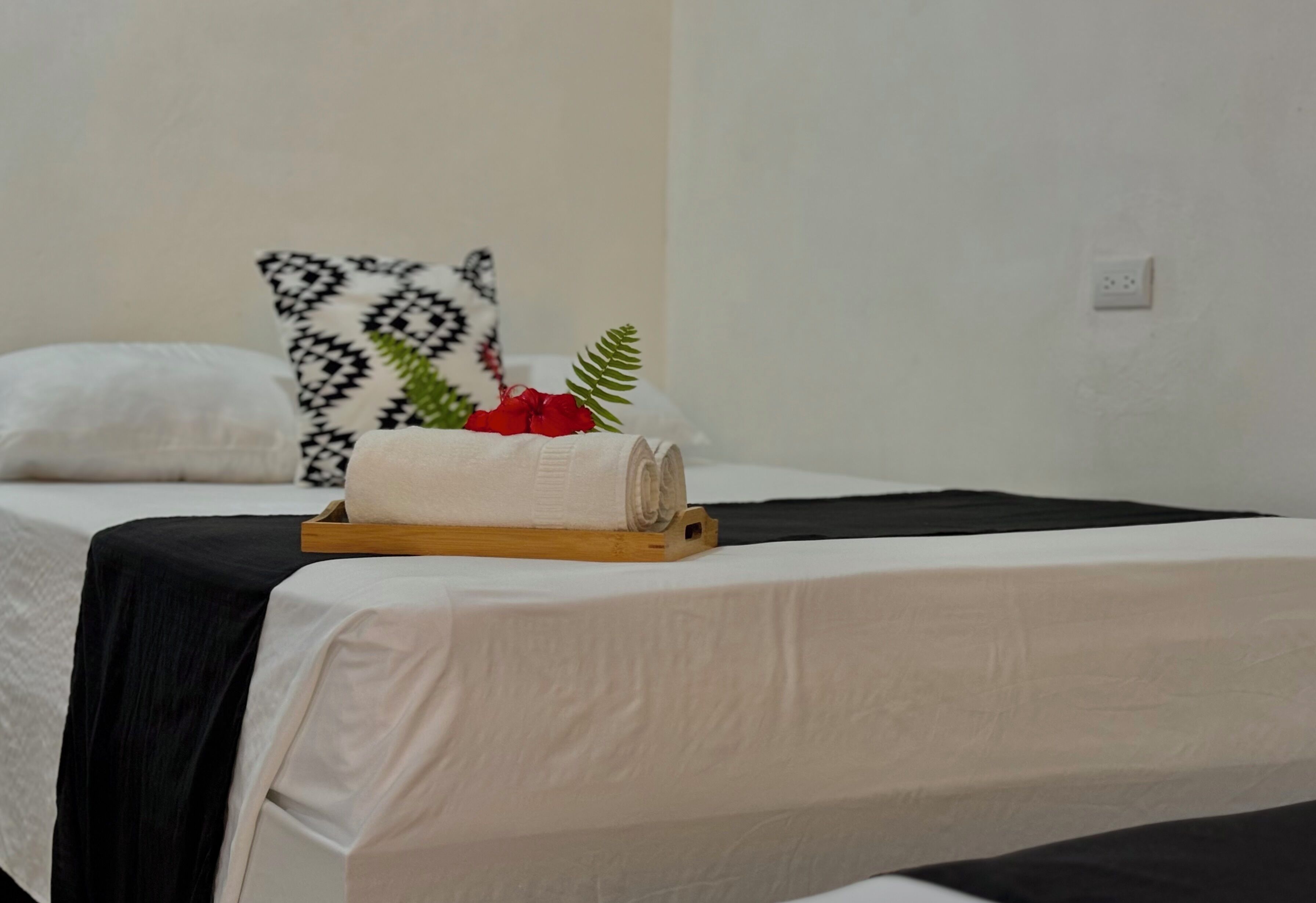 Un lit bien fait, avec du linge de lit blanc, un coussin à motifs noir et blanc et un plateau en bois sur lequel reposent une serviette enroulée et une plante verte.