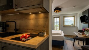 A modern kitchen with a built-in stove, a sink, and a wooden countertop. There is a bed with white bedding in the adjacent room, a dining table with chairs, and a view of the outdoors through the windows. - Appartement 4 "rauschberg" für 2 Personen, 32 m² (Inzell)