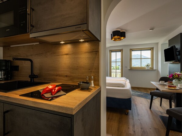 A modern kitchen with a built-in stove, a sink, and a wooden countertop. There is a bed with white bedding in the adjacent room, a dining table with chairs, and a view of the outdoors through the windows.