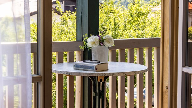 A round table with a book and a vase of flowers on a balcony with a wooden railing, overlooking a mountainous landscape.