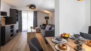 A modern living room with a dining area, featuring a wooden table set for breakfast, a sofa, and a TV.