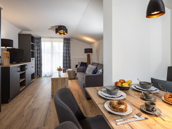 A modern living room with a dining area, featuring a wooden table set for breakfast, a sofa, and a TV.