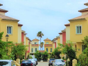 Una hilera de edificios amarillos con detalles rojos y techos de tejas, bordeada por coches aparcados y palmeras.