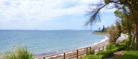 A coastal landscape with a wooden walkway along the beach, leading to a grassy area with trees.