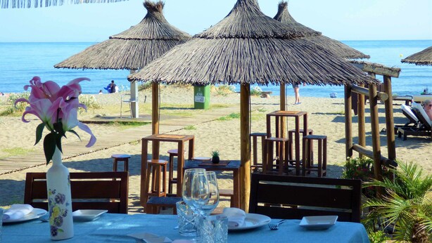 Un restaurante en la playa con sombrillas de paja, mesas y sillas de madera, con vista al mar.