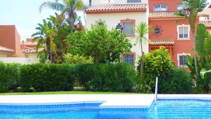Una piscina con borde de azulejos, rodeada de exuberante follaje y palmeras, con edificios al fondo.