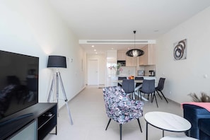 Sala moderna com televisão de tela plana, um tripé e uma poltrona com padrão floral. É visível uma área de jantar com uma cozinha ao fundo.