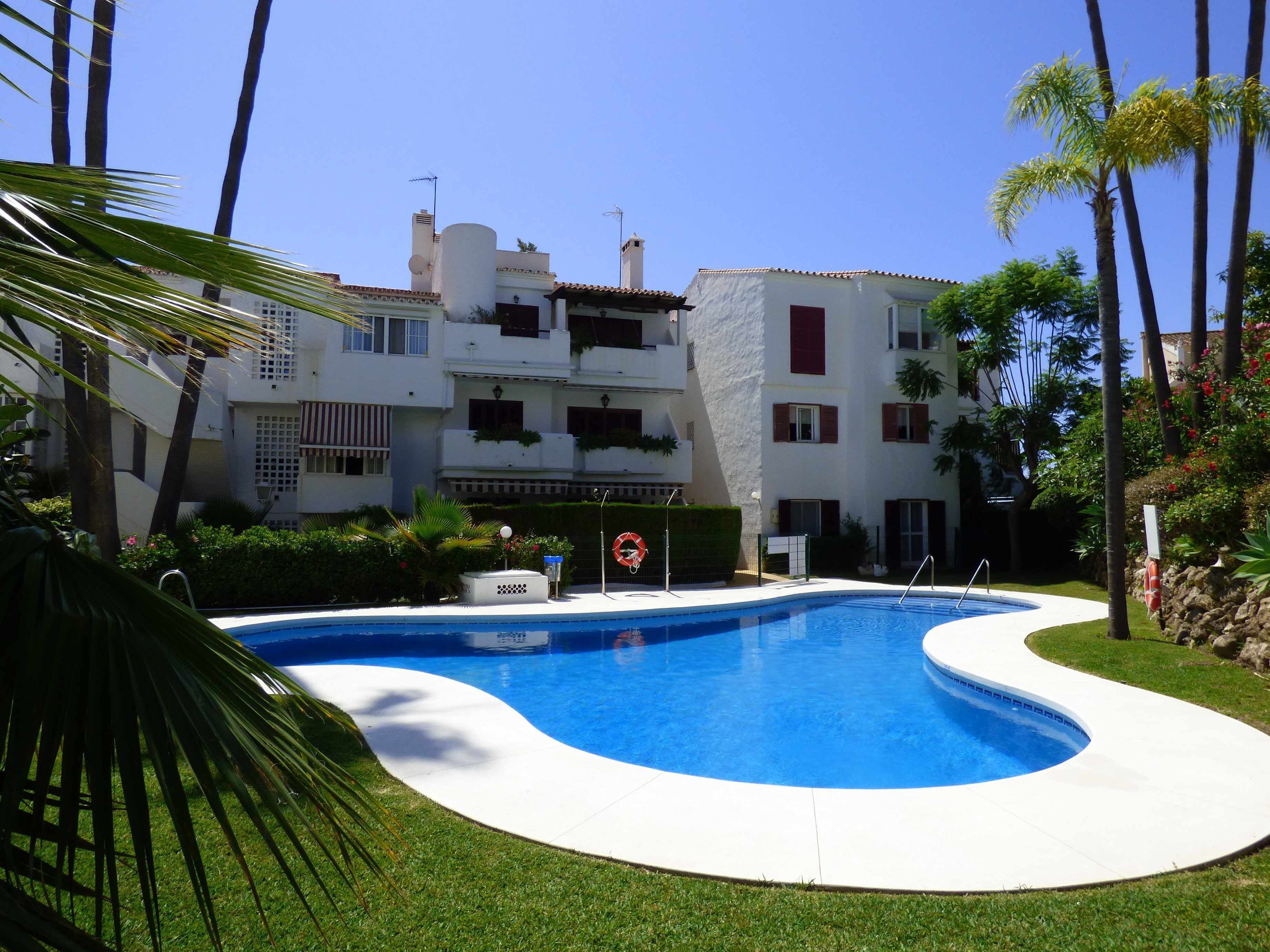 A swimming pool with a white deck, surrounded by palm trees and a building with balconies.