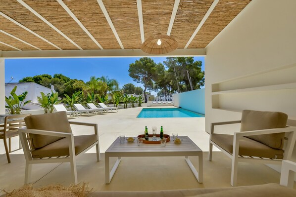 A poolside area with two white lounge chairs, a table with bottles and glasses, and a view of a swimming pool and greenery.