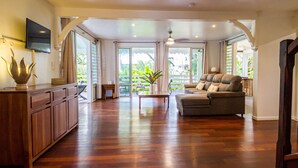 A spacious living area with a wooden floor, a sofa, a television, and a large window offering a view of greenery. - Villa Familiale Avec Piscine Privée à 5 Minutes de la Plage! (Sainte-Rose)
