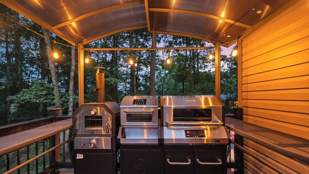 An outdoor kitchen area with a grill, oven, and built-in cabinets, surrounded by a wooden deck and trees.
