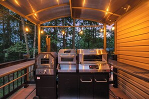 An outdoor kitchen area with a grill, oven, and built-in cabinets, surrounded by a wooden deck and trees. - *Hot Tub*Game Room*Outdoor Kitchen*Bar (State College)