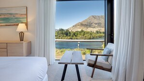 A hotel room with a bed, a small table, a chair, a lamp, and a painting. The room has a large window with a view of a mountain and a body of water. - 3 min to The Remarkables / Lakefront Spa Luxury (Kawarau Falls)