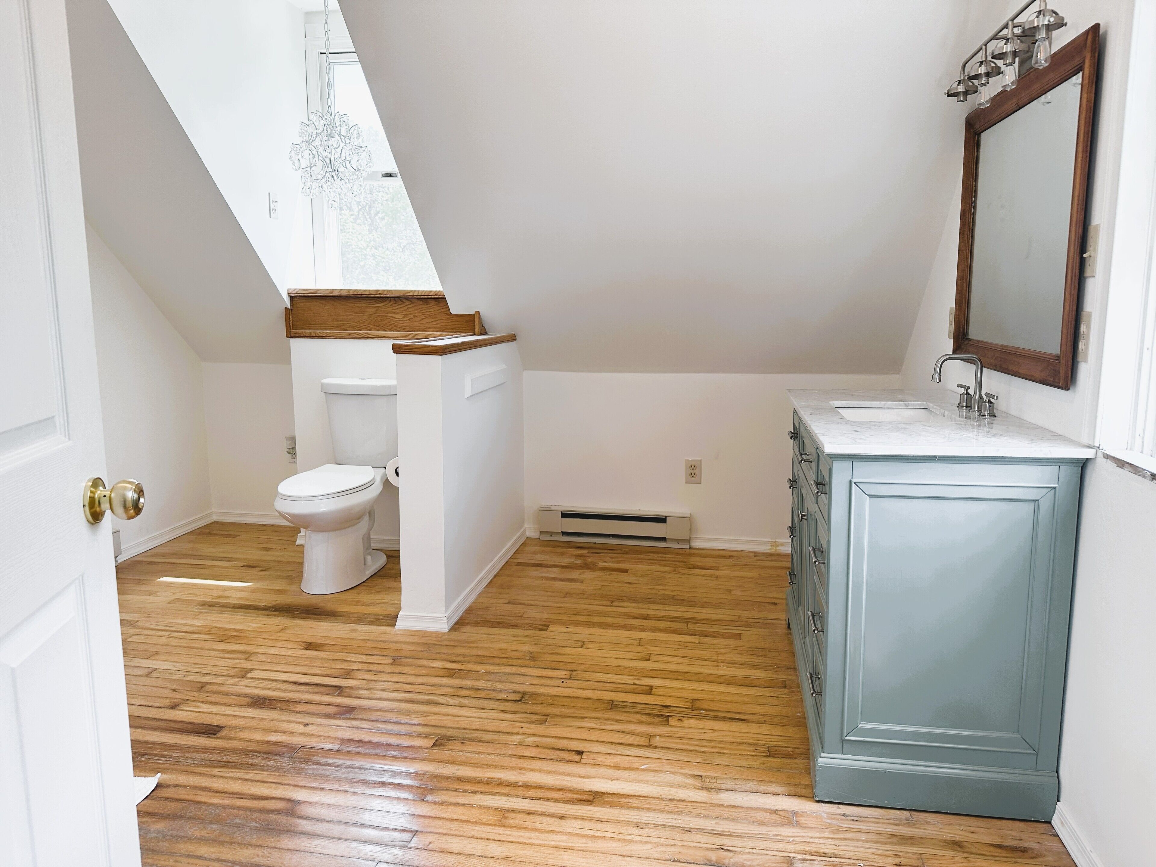 Une salle de bain avec un plancher en bois, des murs blancs, des toilettes blanches et un meuble bleu avec un miroir.