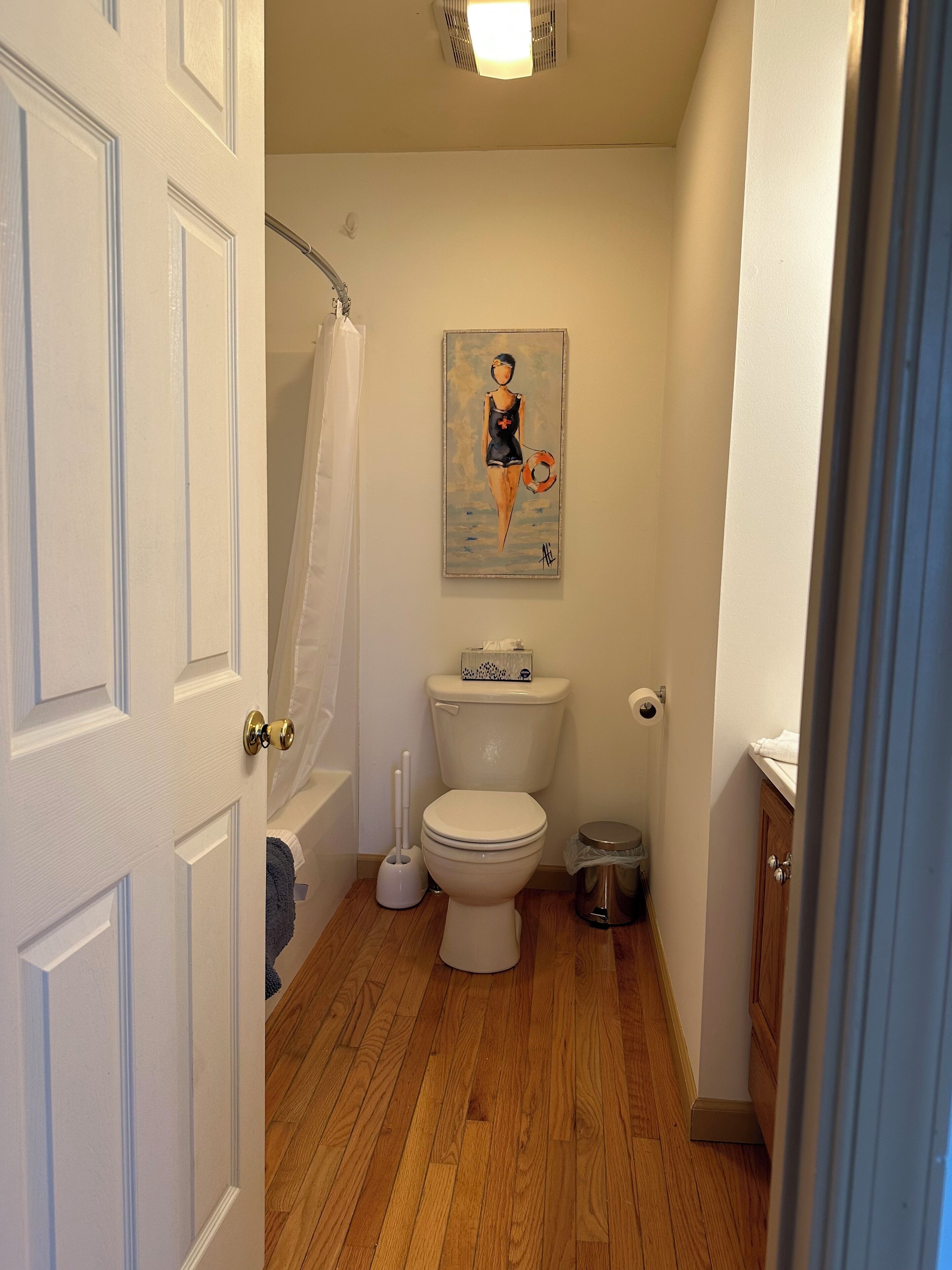 A bathroom with a white toilet, a painting of a lifeguard on the wall, a shower curtain, and a trash can.