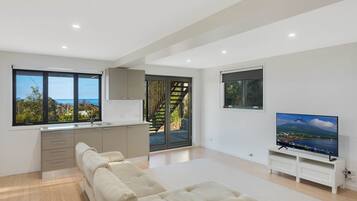 A modern living room with a sofa, a TV, and a kitchen area with a view of the sea.