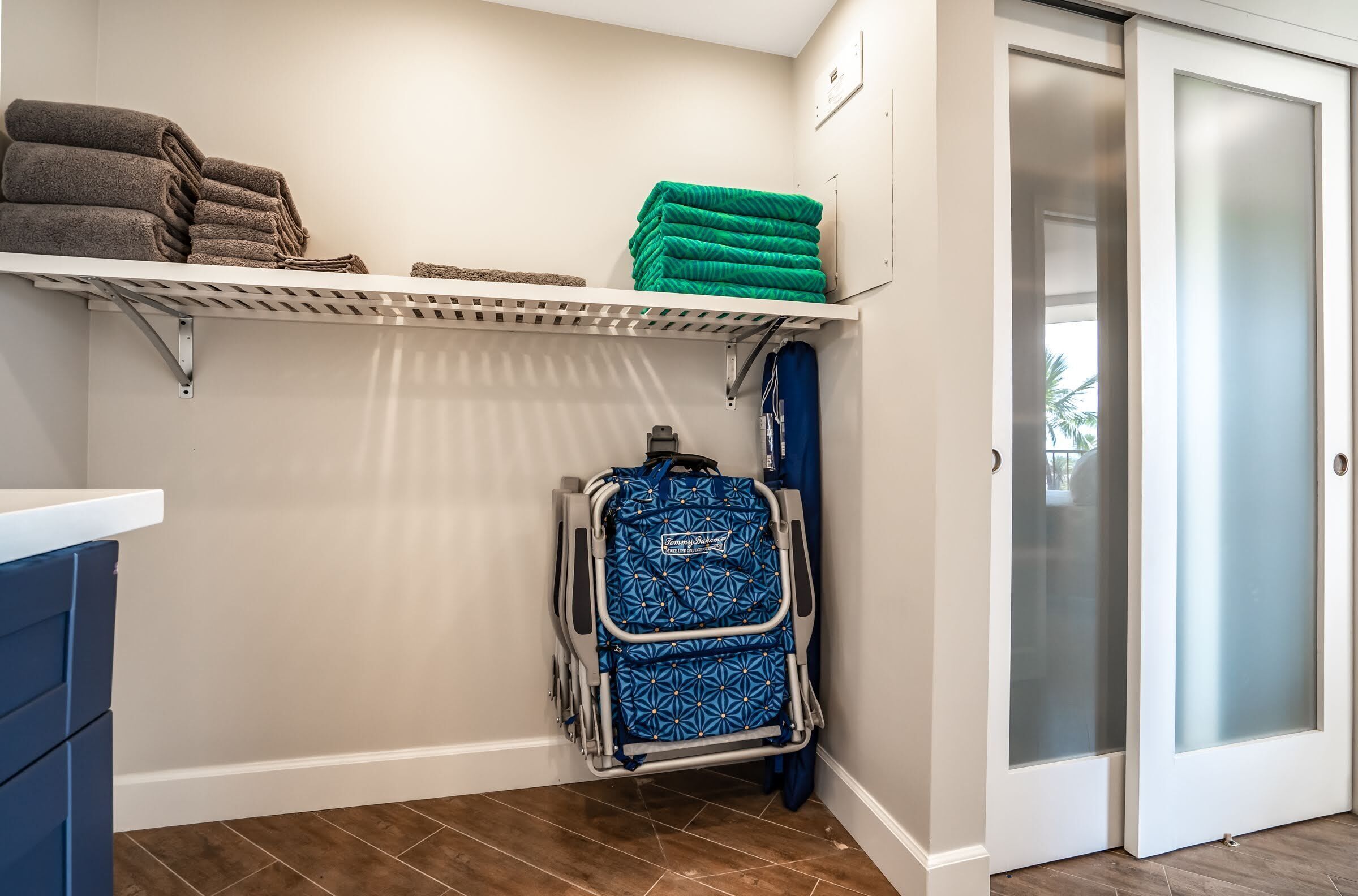 Un baño con un estante blanco lleno de toallas dobladas, una maleta azul y blanca con estampado, y una puerta de cristal satinado.