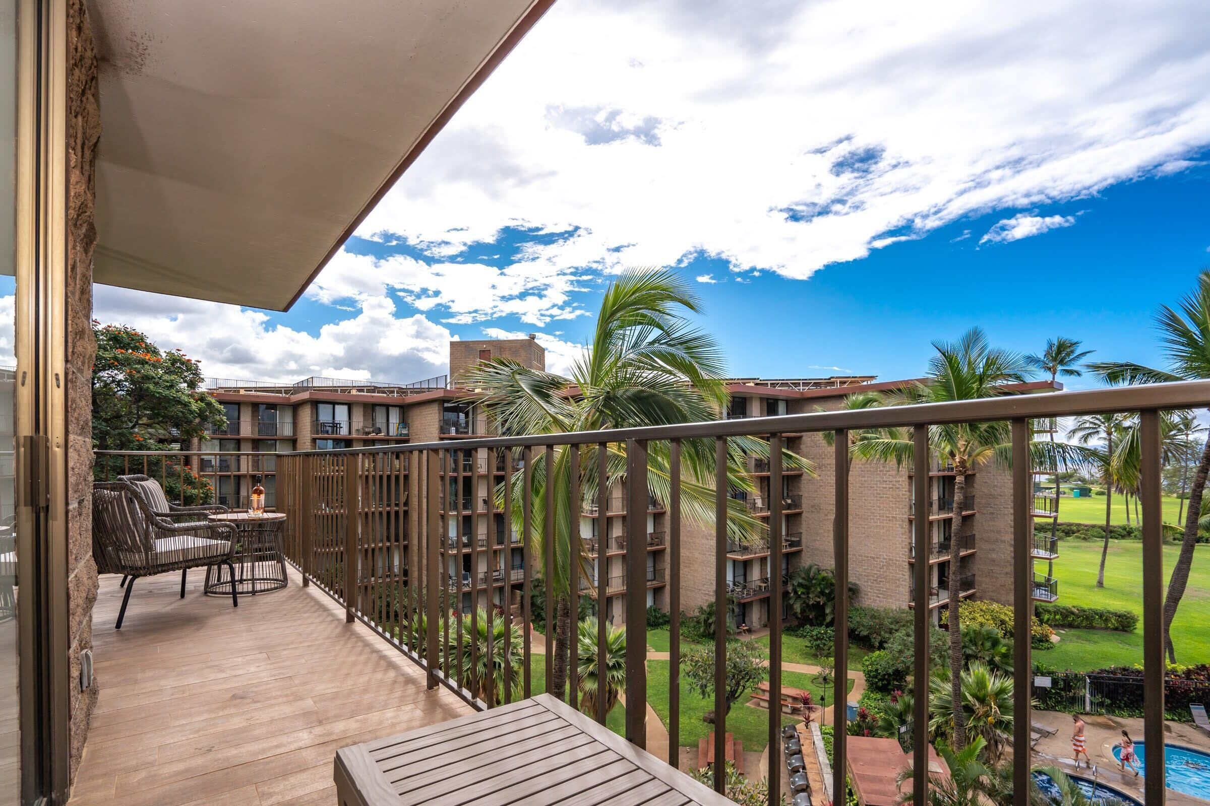 A balcony with wicker furniture, a pool, and a view of a building with multiple balconies and palm trees.