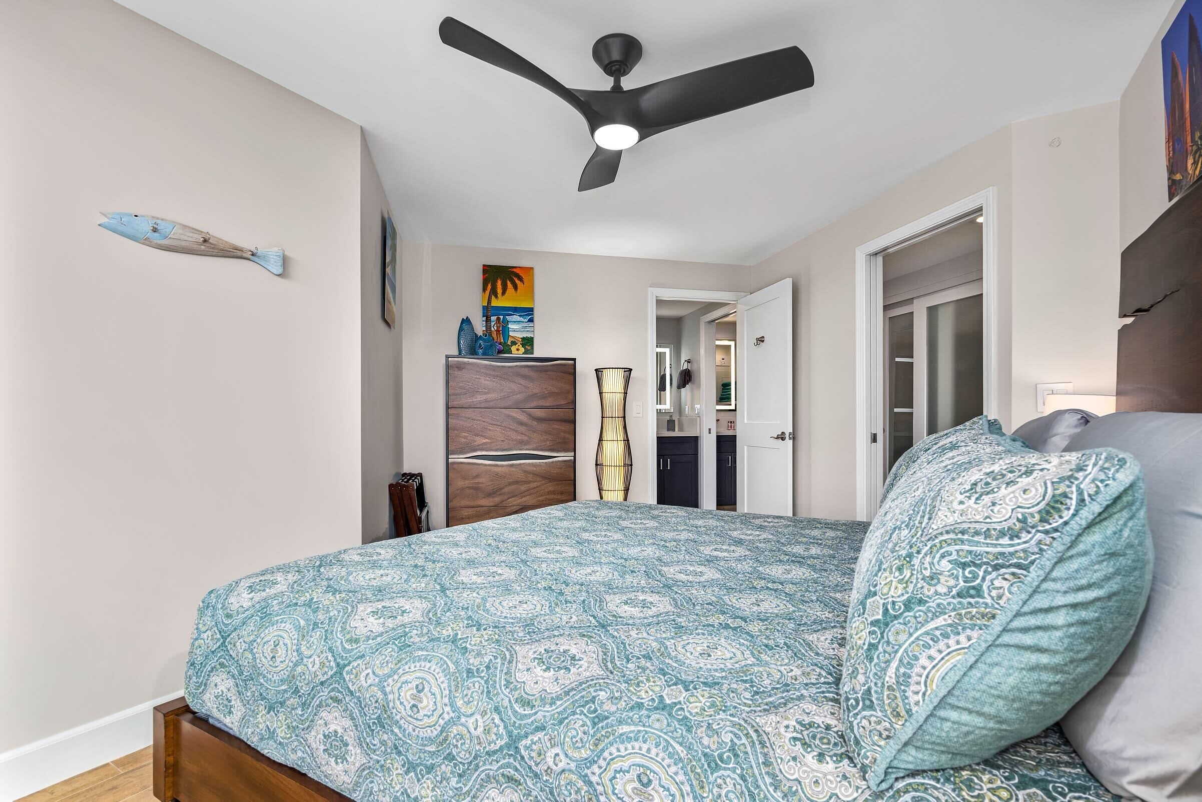 Um quarto com uma cama, um ventilador de teto, um penteou de madeira, um quadro e uma porta que dá para um banheiro.