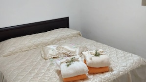 Una cama bien hecha con una colcha acolchada, dos toallas dobladas y un ramita de verdor, además de una mesita de noche con lámpara.