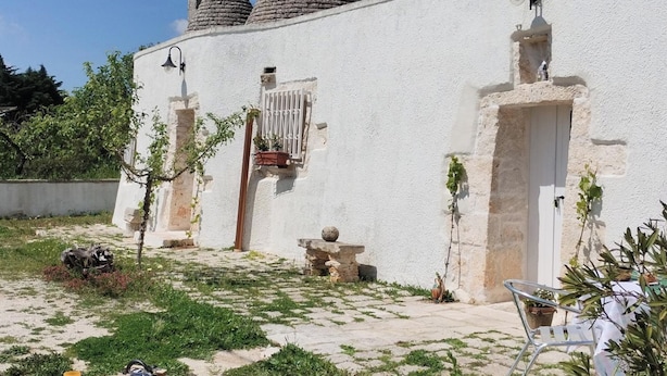 Un edificio blanco con una escalera de piedra que lleva a una puerta, un pequeño jardín con una mesa y sillas, y una planta en maceta.