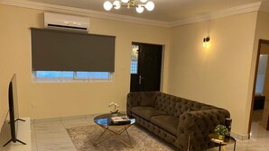 Una sala de estar con un sofá marrón, una mesa de centro de vidrio, una mesita auxiliar con una planta, un televisor de pantalla plana y un equipo de aire acondicionado.
