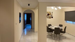 Un comedor con una mesa redonda, sillas y una araña. Hay un televisor montado en la pared y una puerta que da a otra habitación.