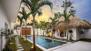 A modern pool area with a clear pool, stone stepping stones, palm trees, and a thatched-roof structure. - Villa Cyma by Alfred in Bali (Seminyak)