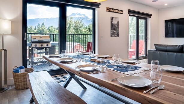 Uma sala de jantar com uma longa mesa de madeira posta para quatro, churrasqueira externa e vista para montanhas através de grandes janelas.