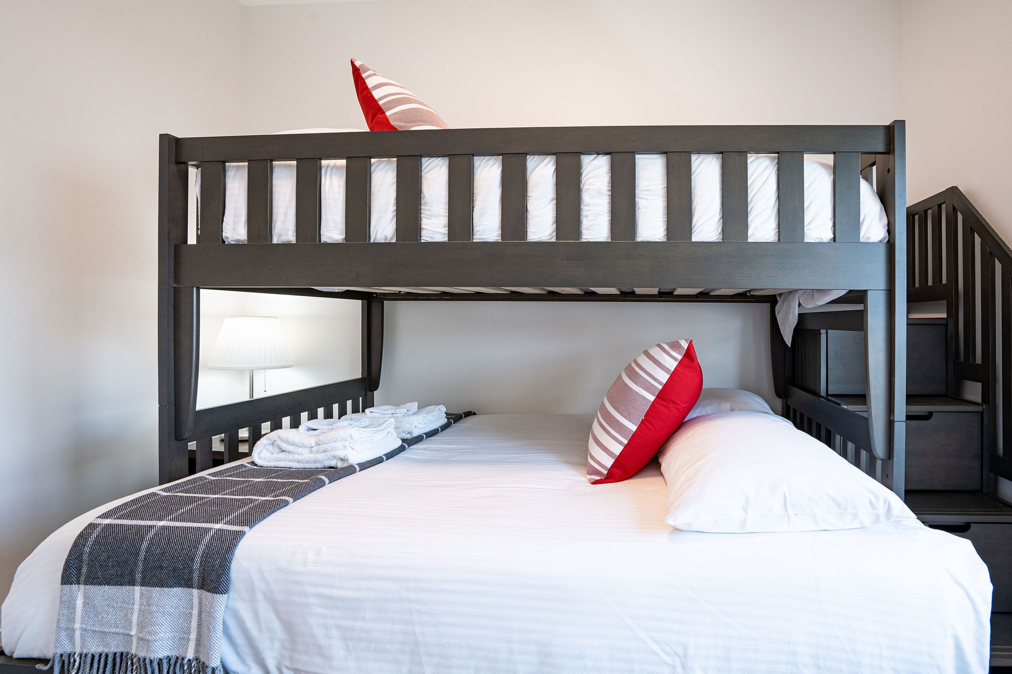 A bunk bed with white bedding and striped pillows, a small table with a lamp, and a staircase to the right.