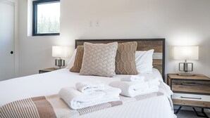 Cama bem arrumada com lençóis brancos e duas almofadas, mesa de cabeceira com abajur, e janela com vista para árvores.