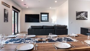 Uma sala de jantar com uma mesa de madeira posta para seis pessoas, um sofá preto e uma janela grande.