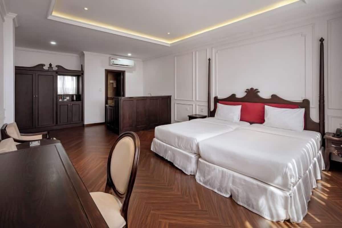A hotel room with a large bed, a wooden desk, a chair, a wardrobe, and an air conditioning unit.