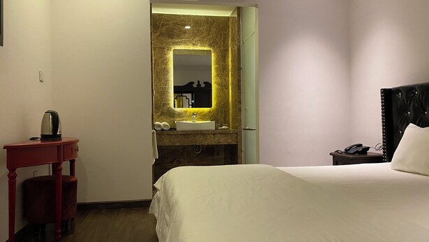 A hotel room with a bed, a vanity with a mirror, a red side table, and a nightstand with a phone. - Mia Hotel (Feliz Hotel Binh Duong) (Ho Chi Minh City)