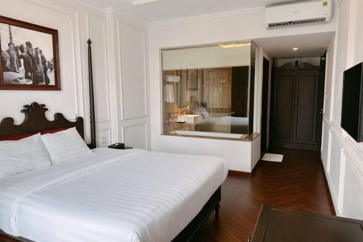 A hotel room with a large bed, a window with a view, a wardrobe, and an air conditioning unit.