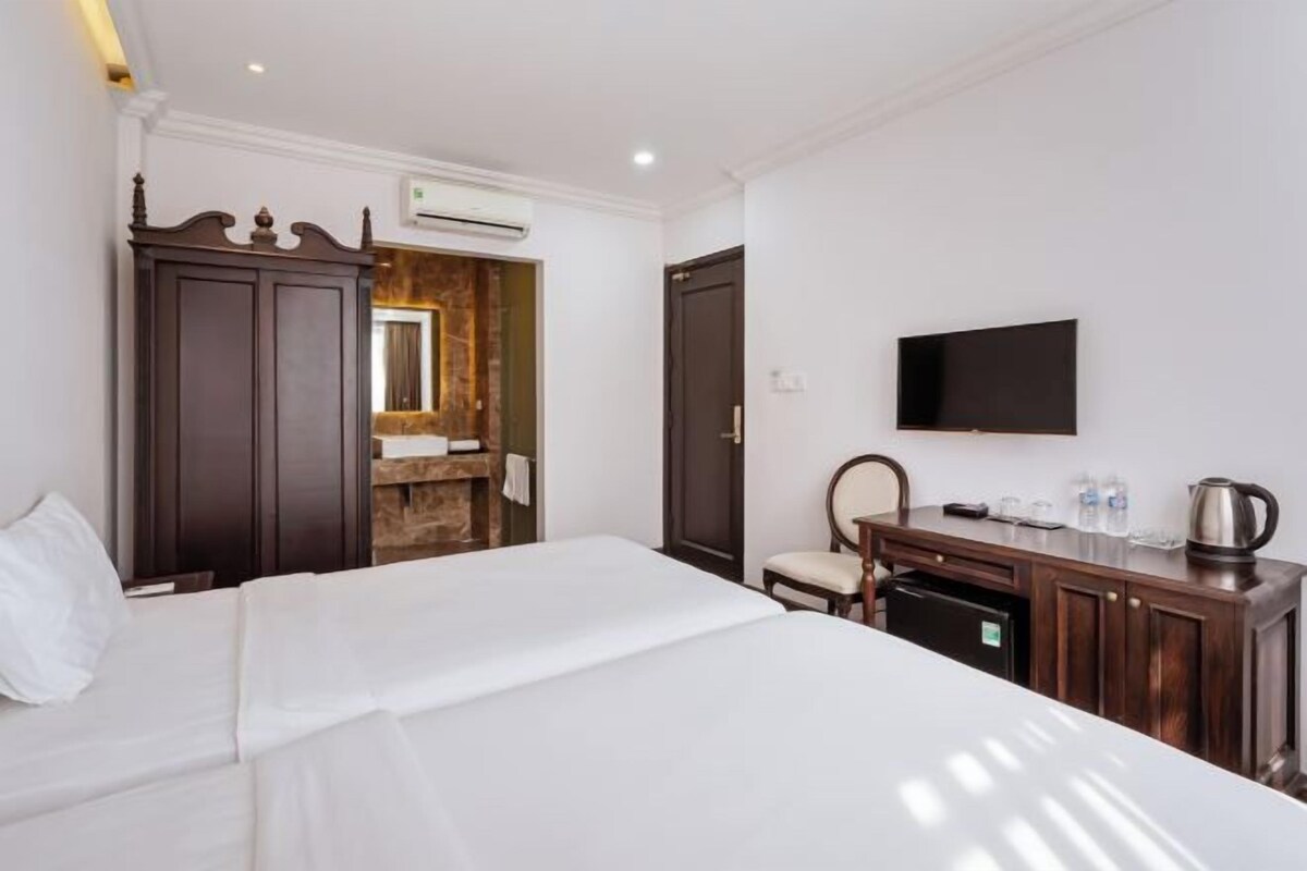 A hotel room with a bed, a wooden wardrobe, a television, a small table with a kettle, and a bathroom door.