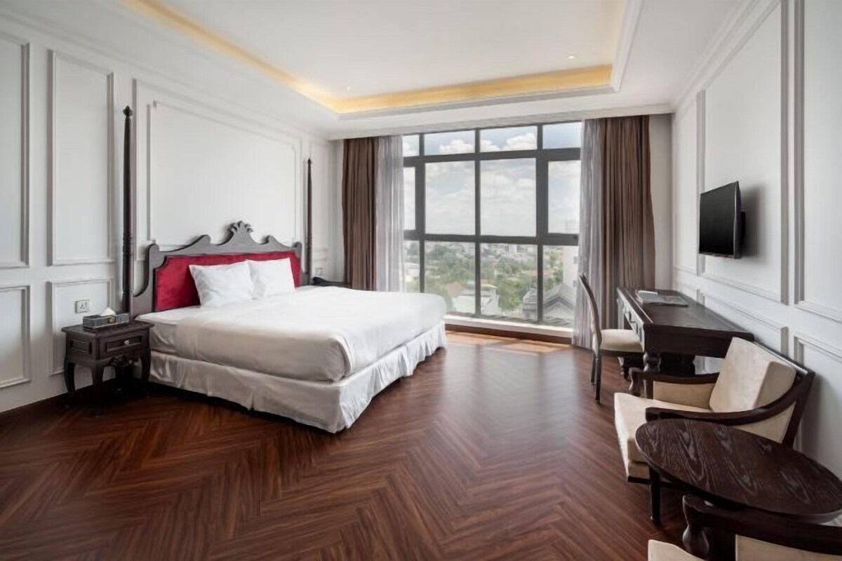 A hotel room with a large bed, a desk, a chair, a TV, and a window with curtains.