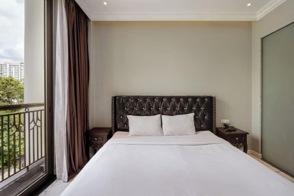 A hotel room with a large bed, a headboard with a tufted design, a nightstand, and a view of the outdoors.