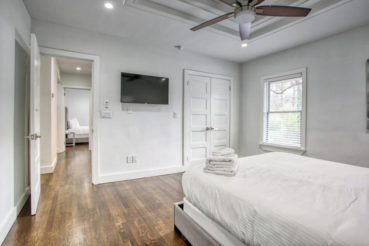 A bedroom with a bed, a ceiling fan, a TV, a door, and a window.