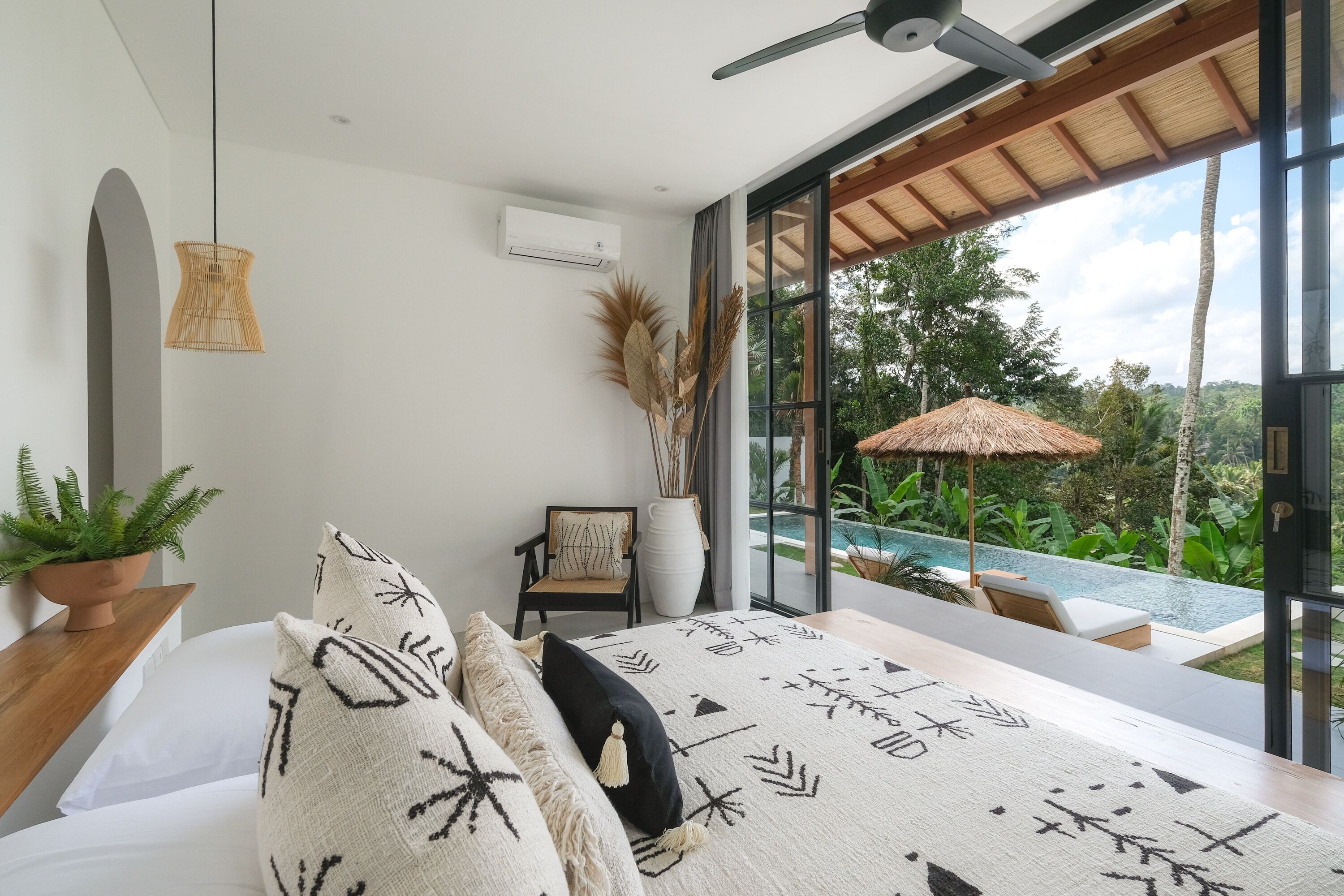 A modern bedroom with a large bed, a ceiling fan, a view of a swimming pool, and a potted plant.