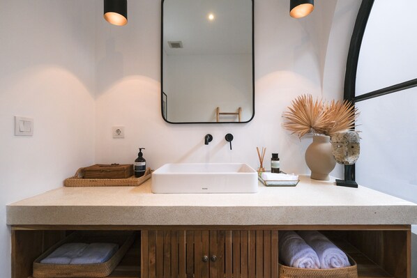 Une salle de bain avec un lavabo blanc, un miroir et un meuble en bois en dessous. On y trouve deux paniers sur le comptoir, une bouteille de savon et un vase contenant des plantes séchées.