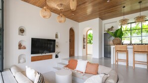A modern living room with a large flat-screen TV, white sofas, and a coffee table. There are wicker pendant lights, a wooden ceiling, and a view of greenery through large windows. - Villa Lestari Ubud (Payangan)